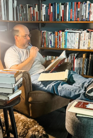 Man reading a book in a cozy armchair surrounded by shelves of books, with a notebook and a dog resting nearby, reflecting a commitment to lifelong learning and personal growth.